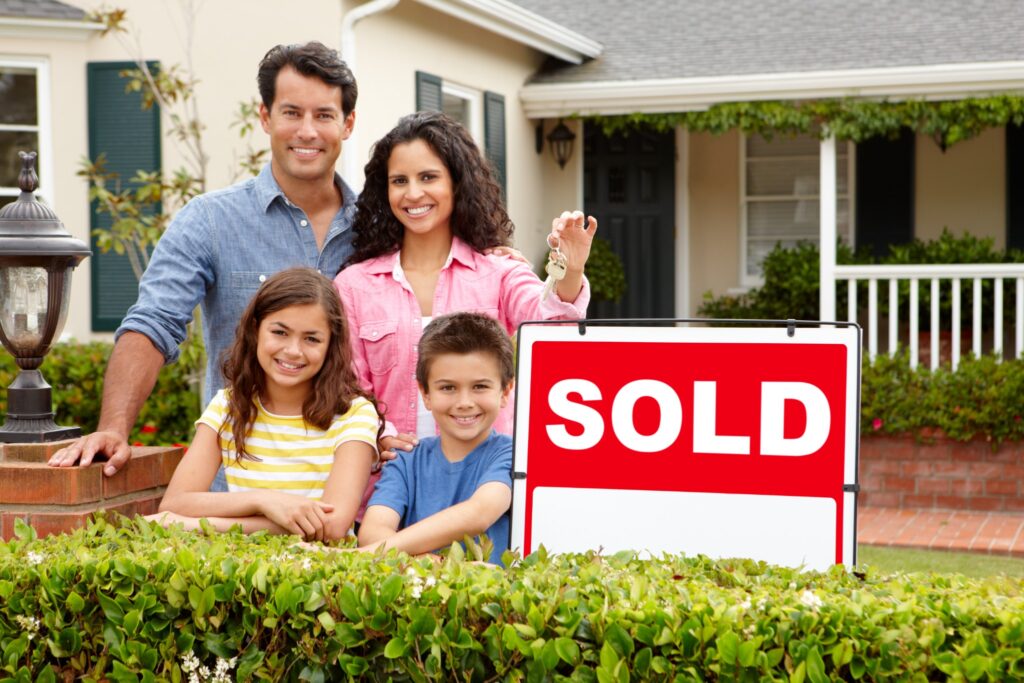 family of two parents and two kids holding keys next to a sold sign in front of their new home