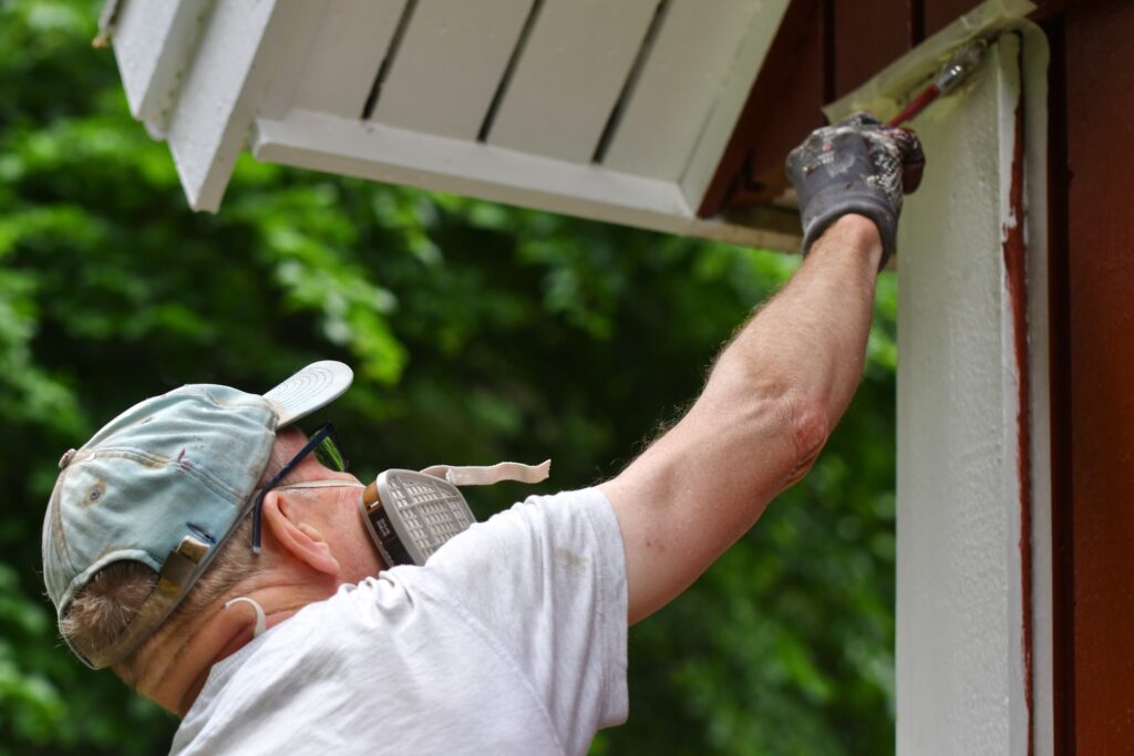 How often do you need to repaint your house? It depends on many factors. Image of man painting the exterior of a house.