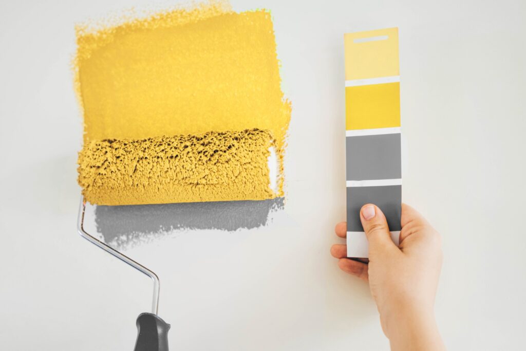 Image of someone painting a wall yellow with a roller while holding a paint swatch in the other hand.